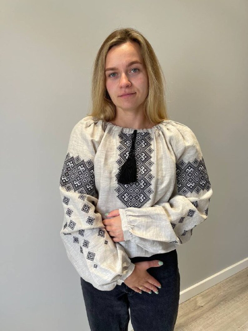 May include: A white linen blouse with black embroidered geometric patterns. The blouse has a gathered neckline and long sleeves with a black tassel.