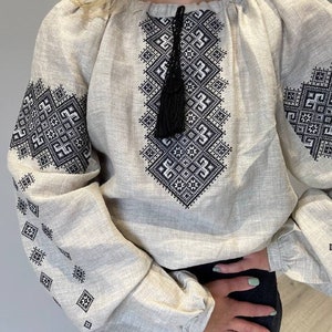 May include: A white linen blouse with black embroidered geometric patterns. The blouse has a gathered neckline and long sleeves with a tassel detail at the neckline.