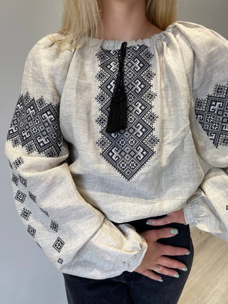 May include: A white linen blouse with black embroidered geometric patterns. The blouse has a gathered neckline and long sleeves with a tassel detail at the neckline.