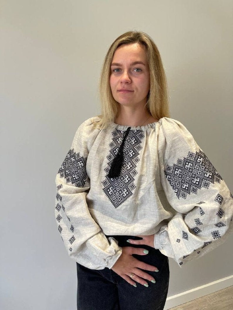 May include: A white linen blouse with black embroidery featuring a geometric pattern. The blouse has a round neckline and long sleeves.