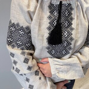 May include: A beige linen blouse with black embroidered geometric patterns and a black tassel. The blouse has long sleeves with ruffles at the cuffs.