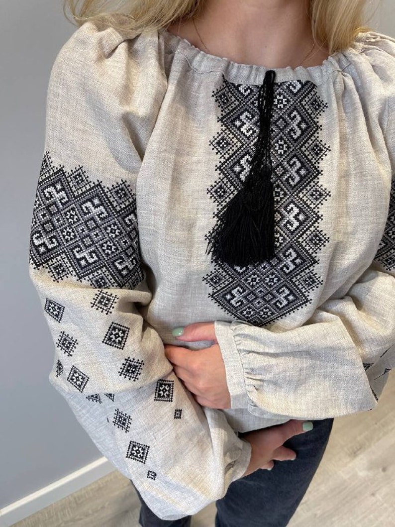 May include: A beige linen blouse with black embroidered geometric patterns and a black tassel. The blouse has long sleeves with ruffles at the cuffs.