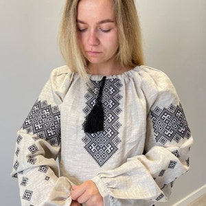 May include: A beige linen blouse with black embroidered geometric patterns and a black tassel. The blouse has long sleeves with gathered cuffs.
