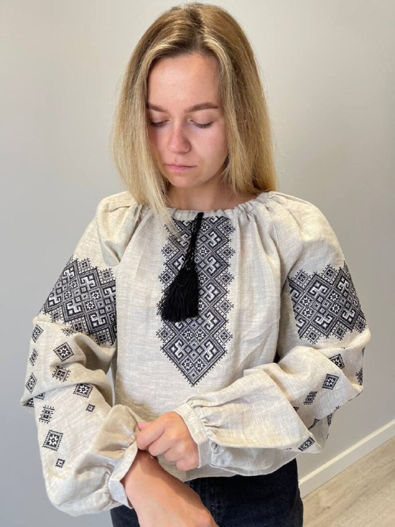 May include: A beige linen blouse with black embroidered geometric patterns and a black tassel. The blouse has long sleeves with gathered cuffs.