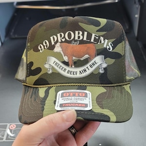 Op de afbeelding: Een camouflage trucker cap met een bruine koe en de tekst "99 Problems but Freeer Beef Ain't One." De pet heeft een zwart mesh achterkant en een wit label met de tekst "OTTO World Famous Foam Front OSFM Trucker Durable Snap Closure".