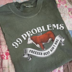 Op de afbeelding: Olijfgroen t-shirt met de tekst "99 Problems but Freezer Beef Ain't One". De afbeelding toont een bruine en witte koe op een spandoek. Het shirt is opgevouwen op een patchwork quilt.
