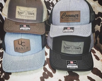 May include: Four different baseball caps with leather patches. The patches have different logos and text, including "Brady Cowan Livestock", "Coomer Contracting", "UC Ranch Properties", and "Brady Cowan Livestock".