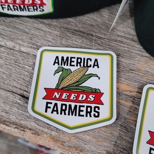 May include: A white sticker with a green border and a yellow inner border. The sticker has a red banner with the text "NEEDS FARMERS" and the text "AMERICA NEEDS FARMERS" above the banner. A cartoon image of an ear of corn is in the center of the sticker.