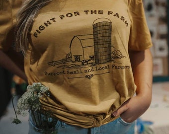Könnte beinhalten: Ein senfgelbes T-Shirt mit einer schwarzen Grafik einer Farm-Szene. Die Grafik zeigt einen Silo, einen Stall und den Text "Fight for the Farm. Support Small and Local Farmers".