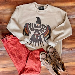 May include: A beige sweatshirt with a graphic of a stylized eagle in black, red, orange, yellow, blue, and green. The sweatshirt is paired with red leggings and brown high-top sneakers.