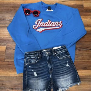 May include: A blue sweatshirt with the word "Indians" in white and red script, paired with distressed denim shorts and red sunglasses. The shorts have a frayed hem.