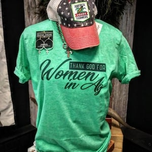 May include: Green t-shirt with the text "Thank God for Women in Ag" in black lettering. The shirt is displayed on a mannequin wearing a red and white baseball cap with the text "America Needs Farmers".