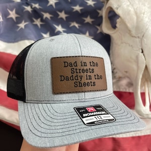 Op de afbeelding: Een grijze en zwarte trucker-pet met een bruin leren patch waarop staat "Dad in the Streets Daddy in the Sheets". De pet heeft een zwart mesh-achterpaneel en een verstelbare Richardson 112-riem.