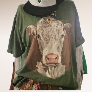 May include: A green t-shirt with a white cow graphic printed on the front. The cow has a brown nose and a gold butterfly charm on its head.