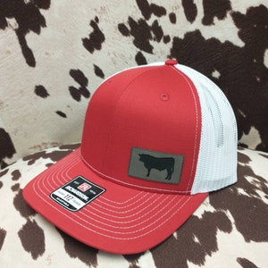 Peut inclure: Casquette de camionneur rouge et blanche avec un patch marron représentant une silhouette noire de vache. La casquette est dotée d'une boucle réglable Richardson 112.