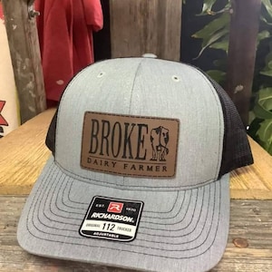 Op de afbeelding: Een grijze en zwarte trucker-pet met een bruin leren patch waarop "Broke Dairy Farmer" staat. De pet is verstelbaar en heeft een Richardson 112 logo.