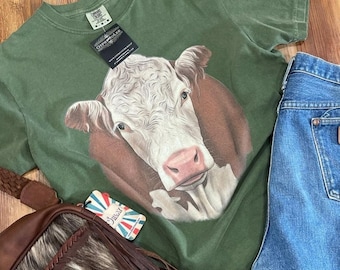 May include: Green short-sleeved t-shirt with a close-up image of a white and brown cow's face. The shirt has a tag that says "Outlaw" and "Cowpoke" on it.