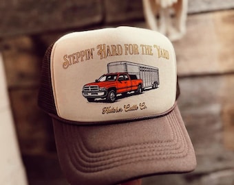 May include: A trucker hat with a brown brim and mesh back. The front panel is tan with the text "Steppin' Hard For The Yard" and an illustration of a red truck pulling a trailer. The text "Hatcher Cattle Co." is below the truck.
