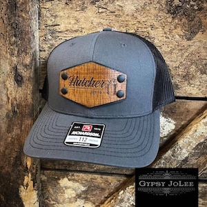 May include: A grey and black trucker hat with a wooden patch that reads "Hatcher Dairy". The hat is adjustable and has a Richardson 112 label.
