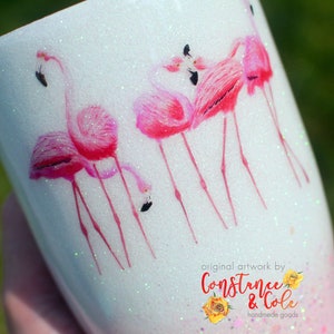 May include: White glitter tumbler with a pink flamingo design. The design features five pink flamingos with black eyes and beaks. The tumbler has the text "original artwork by Constance & Cole handmade goods" at the bottom.