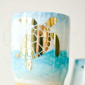May include: A blue and gold tumbler with a sea turtle design. The tumbler has a white base and a gold glitter bottom.