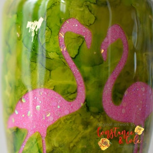 May include: Green and pink tumbler with a glittery pink flamingo design. The tumbler has a green background with a watercolor effect. The text "original artwork by Constance & Cole handmade goods" is at the bottom of the tumbler.