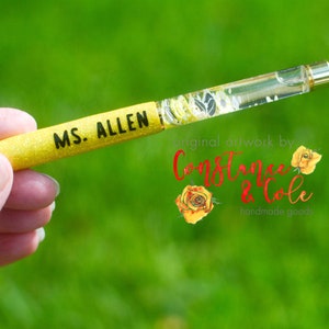 May include: Yellow glitter pen with a clear resin section containing dried flowers. The pen is personalised with the text "MS. ALLEN". The pen is held in a hand against a green background. The text "original artwork by Constance & Cole handmade goods" is visible in the background.