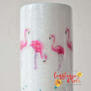 May include: A white glitter tumbler featuring a watercolor design of pink flamingos. The tumbler has a cylindrical shape and is decorated with a repeating pattern of the birds. The text "original artwork by Constance & Cole handmade goods" is visible.