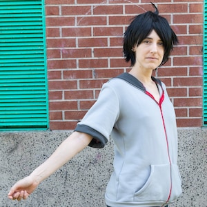 May include: A person wearing a grey zip-up hoodie with a red zipper and black trim on the sleeves. The person is wearing dark blue jeans and has short black hair.