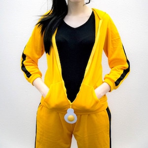 May include: A yellow tracksuit with black stripes on the sleeves and legs. The jacket is zipped up and has a hood and pockets. The zipper pull is a fried egg design. The person is wearing a black v-neck shirt.