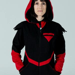 Puede incluir: Una sudadera con capucha negra con detalles rojos. La sudadera tiene una cremallera y un parche triangular en el pecho. Las mangas y la parte inferior de la sudadera son rojas.
