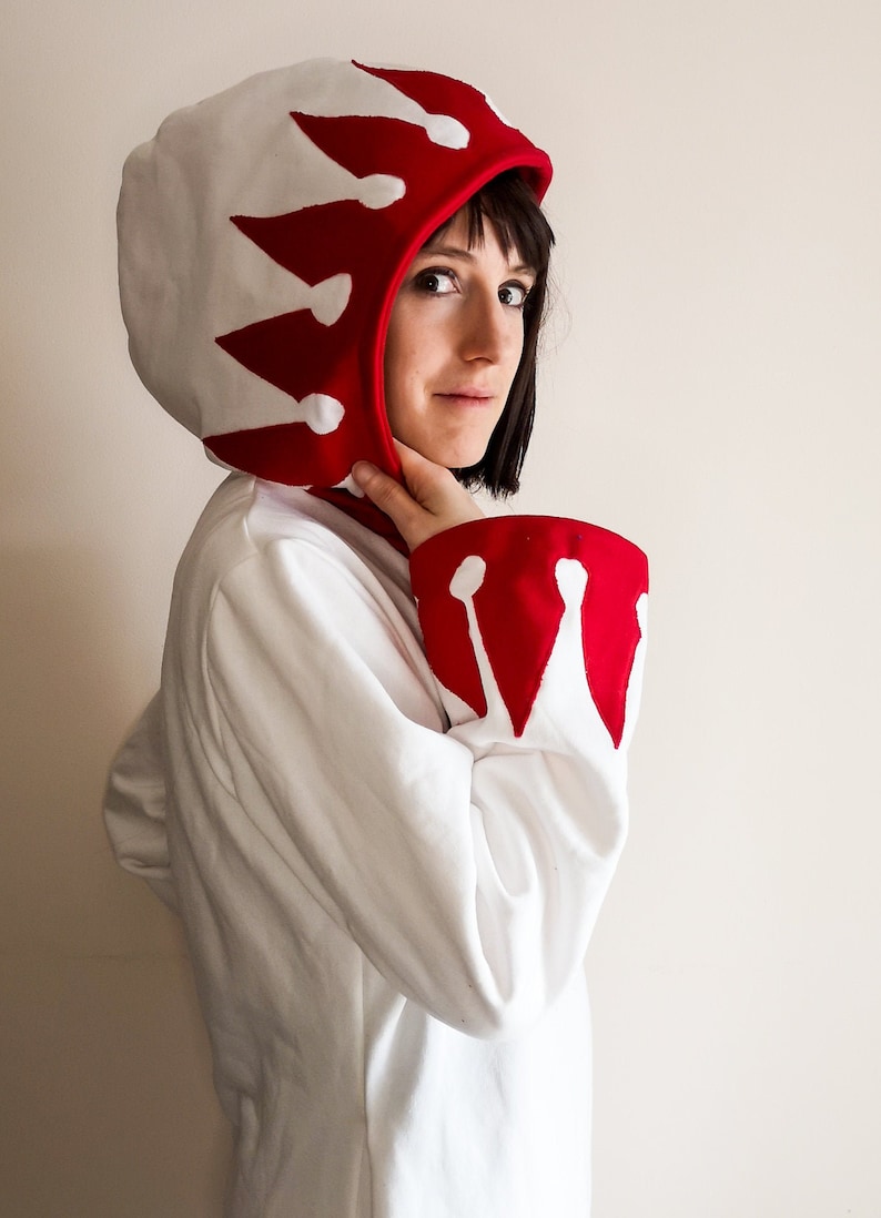 May include: White hooded sweatshirt with red crown design on the hood and sleeves. The hood is pulled up over the head.