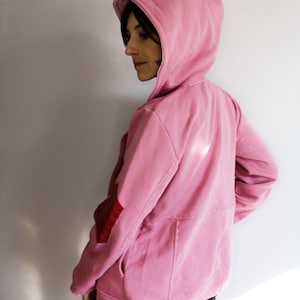 May include: A light pink hooded sweatshirt with a kangaroo pocket and a red elbow patch. The sweatshirt has a drawstring hood and visible stitching details. The model is wearing black pants.