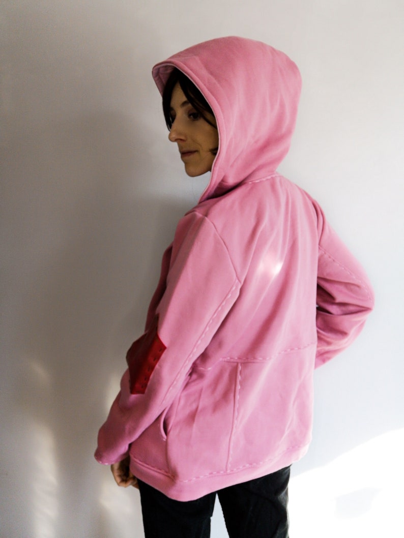May include: A light pink hooded sweatshirt with a kangaroo pocket and a red elbow patch. The sweatshirt has a drawstring hood and visible stitching details. The model is wearing black pants.