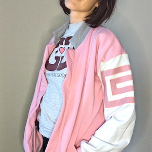 Peut inclure: Une veste bomber rose et blanche avec une fermeture éclair grise et un col gris. La veste a une bande blanche sur la manche avec un motif géométrique rose et blanc. La veste a une poche sur le côté gauche. La veste a une doublure intérieure grise. La veste a un graphique gris et brun sur le devant qui dit "Cheers for Good".