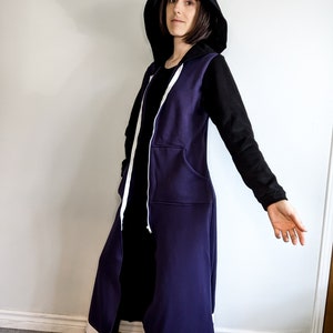 May include: A long, purple hooded sweatshirt with white trim and a black zip. The hood is pulled up and the sweatshirt is worn over a black shirt.