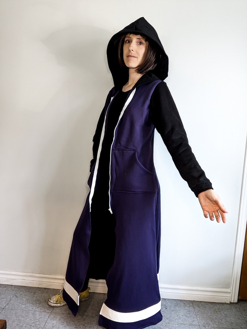 May include: A long, purple hooded sweatshirt with white trim and a black zip. The hood is pulled up and the sweatshirt is worn over a black shirt.
