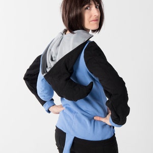 May include: A blue, black, and gray hooded sweatshirt with a unique design. The sweatshirt features black sleeves, a blue body, and a gray hood. A blue tail-like extension hangs from the back. The model is wearing black pants.