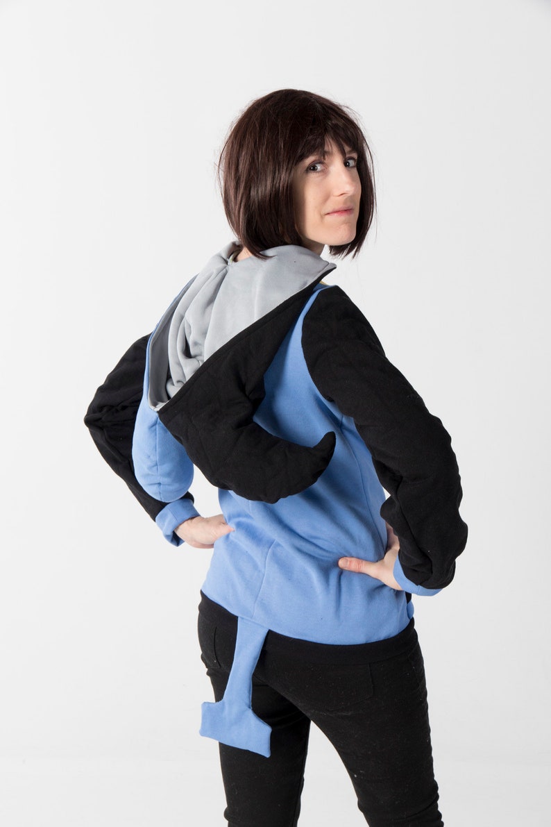 May include: A blue, black, and gray hooded sweatshirt with a unique design. The sweatshirt features black sleeves, a blue body, and a gray hood. A blue tail-like extension hangs from the back. The model is wearing black pants.