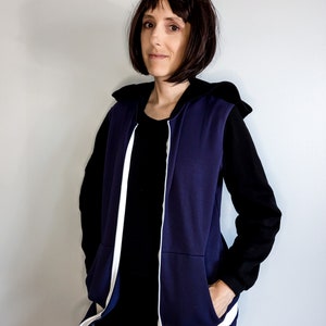 May include: A navy blue hooded vest with white piping and a zip closure. The vest is worn over a black long-sleeved shirt and black trousers.