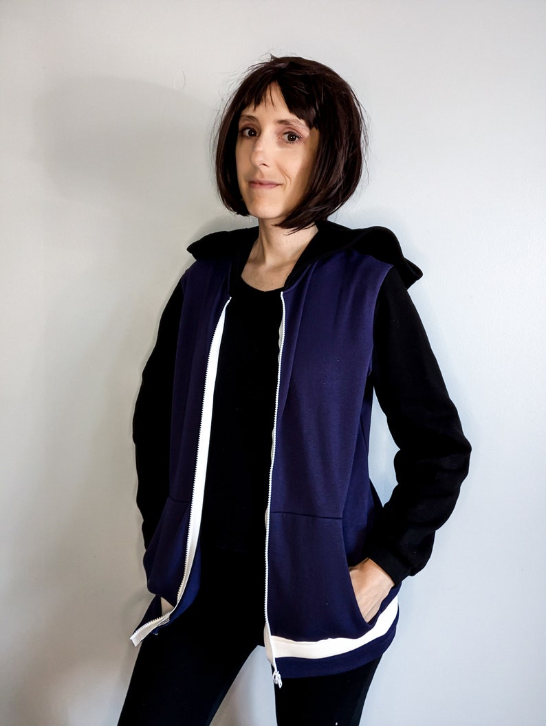 May include: A navy blue hooded vest with white piping and a zip closure. The vest is worn over a black long-sleeved shirt and black trousers.