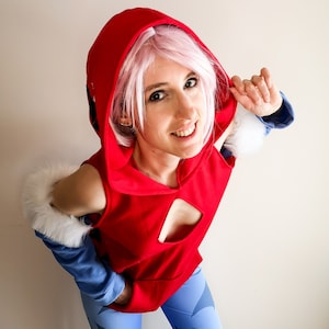 May include: A woman wearing a red hooded top with a white fur trim, blue pants with a geometric pattern, and pink hair.