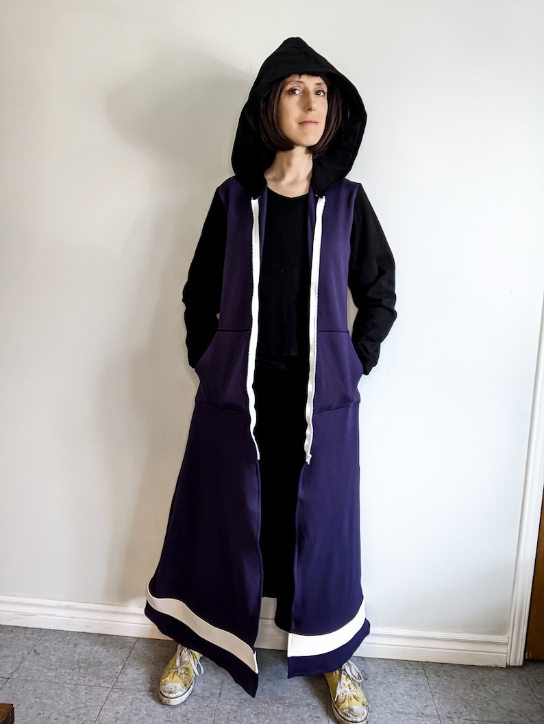 May include: A long purple hooded sweatshirt with white trim and a black zipper. The hood is pulled up and the sweatshirt is unzipped.