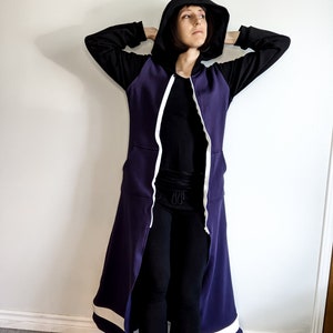 May include: A long purple hooded sweatshirt with white trim and a zip down the front. The hood is pulled up and the sweatshirt is worn with black leggings.