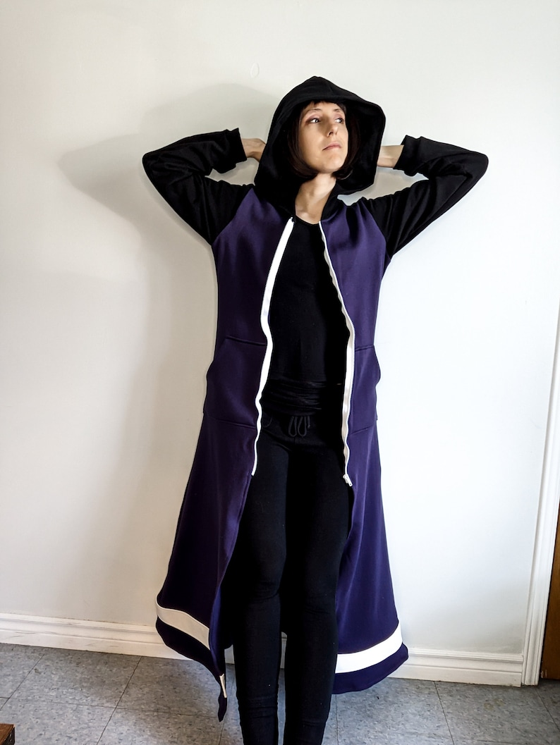 May include: A long purple hooded sweatshirt with white trim and a zip down the front. The hood is pulled up and the sweatshirt is worn with black leggings.