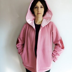 May include: A pink hooded jacket with a satin lining and elbow patches. The jacket has a relaxed fit with pockets and a button detail on the sleeve cuffs. The model is wearing a black shirt underneath.