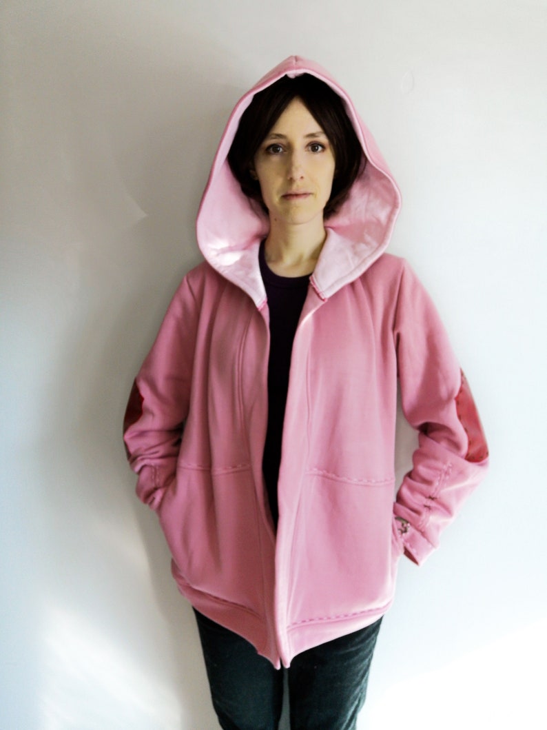 May include: A pink hooded jacket with a satin lining and elbow patches. The jacket has a relaxed fit with pockets and a button detail on the sleeve cuffs. The model is wearing a black shirt underneath.