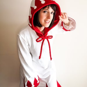 May include: A white hooded robe with red trim and a crown design. The hood is tied with a red ribbon. The robe has a white and red design on the sleeves and hem.