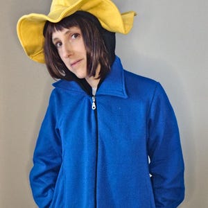 Könnte beinhalten: Eine Person trägt eine blaue Jacke mit Reißverschluss und einen gelben spitzen Hut. Der Hut hat eine breite Krempe und ist aus Filz.