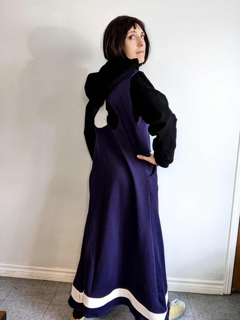 May include: A woman wearing a long, dark purple dress with a white trim at the bottom. The dress has a white heart shape on the back. She is also wearing a black hooded sweatshirt.